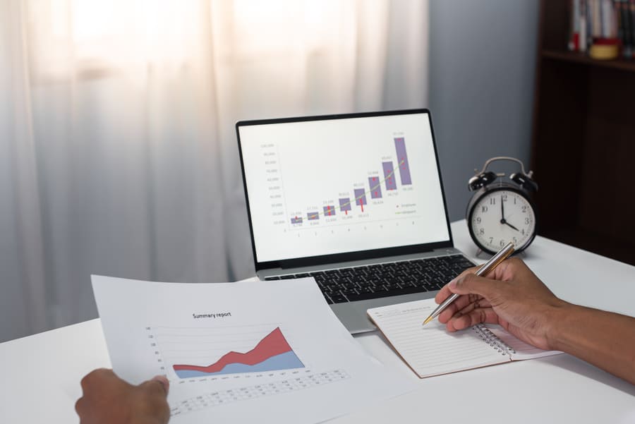 Hands holding a printed summary report with an area chart, next to a laptop showing a rising bar chart and an alarm clock, illustrating the week-ahead market outlook.