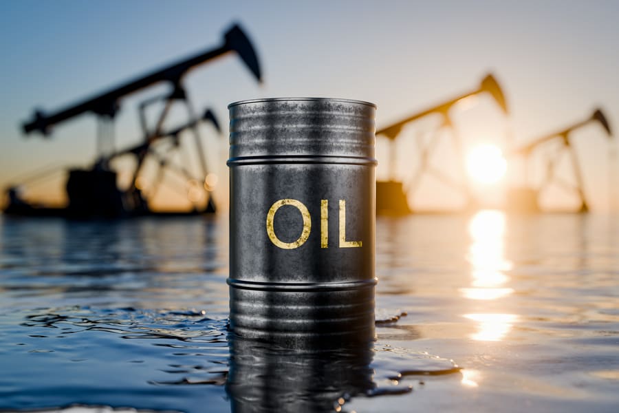 Tong minyak hitam bertulis OIL di atas permukaan minyak berkilat, dengan siluet pam angguk telaga minyak berlatarbelakangkan matahari terbenam