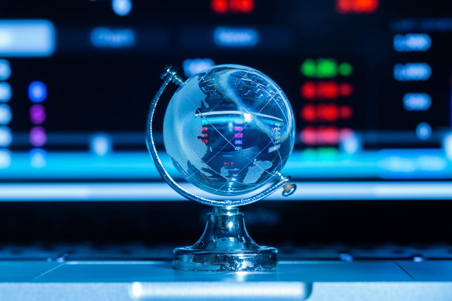 Crystal glass globe on a silver stand resting on a laptop, with a blurred trading screen of red and green market prices in the background