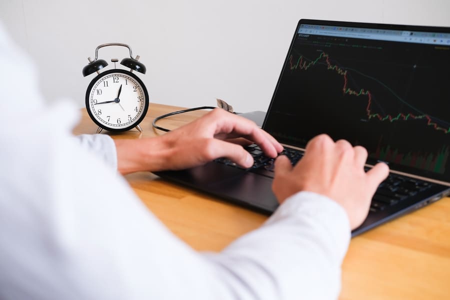 Person typing on a laptop showing a candlestick trading chart, with a black alarm clock on a wooden desk